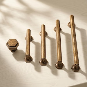 Peut inclure: Un ensemble de cinq pièces de quincaillerie pour armoires en laiton antique, comprenant un bouton hexagonal et quatre poignées barre de longueurs variées. Chaque pièce présente une finition texturée et vieillie avec une forme hexagonale.