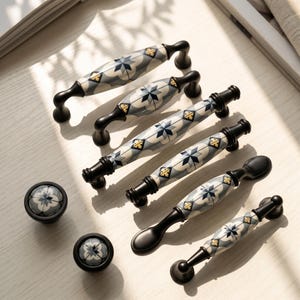 May include: A collection of cabinet hardware including four elongated drawer pulls and two round knobs. Each piece features a ceramic center with a gray, white, and blue star pattern and gold accents, finished with matte black metal ends.