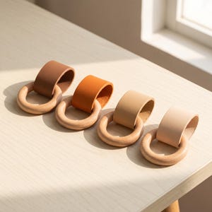 May include: Four teething rings, each consisting of a smooth, light-coloured wooden ring looped through a folded strip of leather. The leather strips vary in colour from dark brown to medium brown, light tan, and a very pale cream or beige shade.