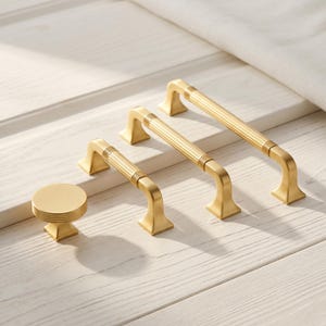 Op de afbeelding: Een set van vier goudkleurige meubelbeslagstukken, waaronder één ronde knop en drie gebogen ladehandgrepen van verschillende lengtes. Elk stuk is voorzien van een geribbelde, cilindrische greep en een vierkante montagebeugel.