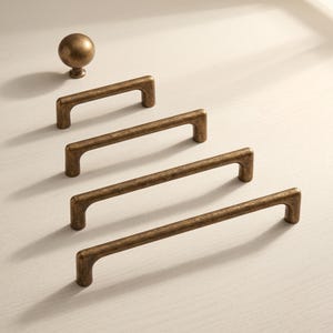 May include: A collection of antique brass hardware including one round cabinet knob and four rectangular drawer pulls of varying lengths. Each piece features a textured, weathered metallic finish with rounded edges and a sturdy, solid construction.