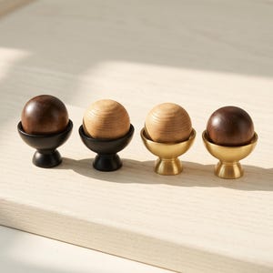 May include: Four cabinet knobs, each featuring a spherical wooden top set into a metal base. Two knobs have dark brown wood spheres on matte black bases, while the other two have light tan wood spheres on brushed gold-toned metal bases.