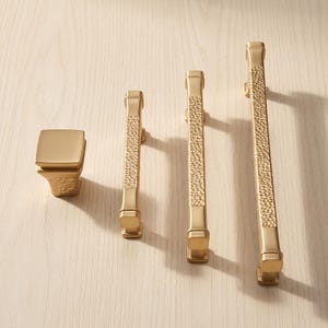 May include: A set of four gold-toned cabinet hardware pieces, including one square knob and three linear pulls of varying lengths. Each piece features a smooth finish on the ends and a textured, hammered-style center section in a metallic gold.