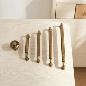 May include: A collection of five antique brass cabinet hardware pieces, including one round knob and four bar pulls of varying lengths. Each piece features a textured, weathered finish with a hexagonal central bar and curved mounting posts.