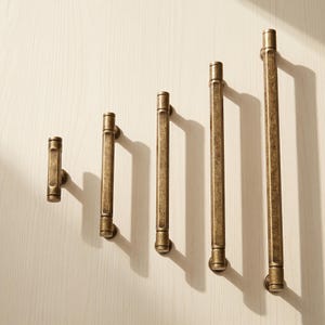 May include: Five antique brass cabinet pulls of varying lengths arranged in a row. Each handle features a hexagonal bar design with a textured, weathered finish and cylindrical end caps, providing a consistent look across different sizes.