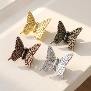 May include: Four butterfly-shaped cabinet knobs in different metallic finishes. One knob is polished gold, one is antique bronze, one is copper, and one is silver. Each knob features a detailed, raised wing pattern with a central body segment.