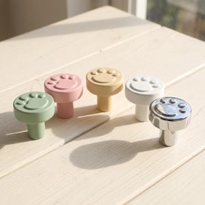 May include: Five circular cabinet knobs featuring a raised paw print design on the top surface. The knobs are finished in matte sage green, matte pink, matte yellow, matte white, and a polished metallic silver finish, each with a cylindrical base.