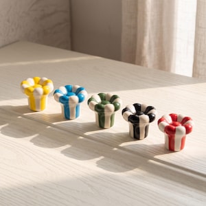 Puede incluir: Cinco pomos de cerámica para armarios con forma de flor y bordes festoneados. Cada pomo presenta un patrón de color distinto: rayas verticales amarillas y blancas, azules y blancas, verdes y blancas, negras y blancas, y rojas y blancas.