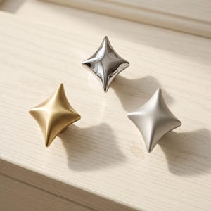 Può includere: Tre pomelli per armadietto a forma di stella con finiture diverse. Un pomello ha una finitura cromata lucida, uno una finitura oro spazzolato e uno una finitura argento opaco. Ogni pomello presenta quattro punte e una superficie centrale curva.