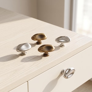 May include: A collection of five cabinet knobs with irregular, organic, pebble-like shapes. Three knobs feature a polished silver-tone metallic finish, while two knobs have a brushed, antique brass-tone finish, each with a central mounting post.