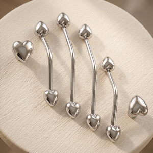 Puede incluir: Un conjunto de herrajes de metal en tono plateado que consta de dos pomos en forma de corazón y cuatro tiradores de cajón curvos, cada uno con extremos en forma de corazón con un acabado pulido y reflectante y un diseño suave.