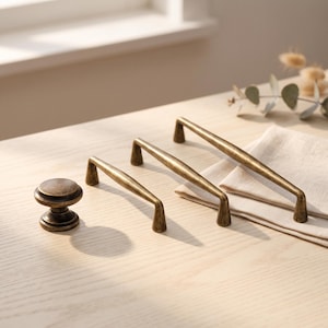 May include: A set of antique brass hardware including one round cabinet knob and three arch-shaped drawer pulls of varying lengths. Each piece features a smooth, metallic finish with subtle dark distressing, showcasing a classic design.
