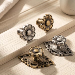 Puede incluir: Cuatro pomos de metal para armarios con acabados en latón antiguo y plata. Dos pomos tienen un diseño sencillo en forma de flor, mientras que los otros dos están montados sobre una placa decorativa rectangular adornada con volutas.