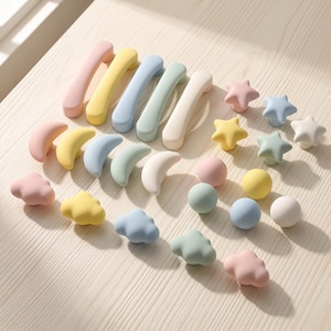 May include: A collection of cabinet hardware in pastel colors including pink, yellow, blue, green, and white. The set features five arched handles, five crescent moon knobs, five cloud-shaped knobs, five star-shaped knobs, and five round ball knobs.