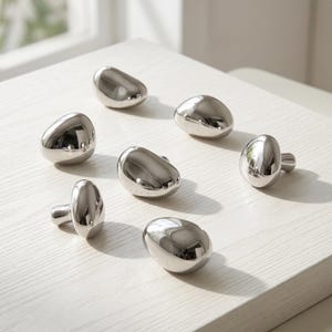 Puede incluir: Un conjunto de seis pomos para armarios en tono plateado, cada uno con una forma suave e irregular similar a un guijarro, con un acabado metálico pulido altamente reflectante. Estas piezas de herraje presentan una forma orgánica y redondeada.