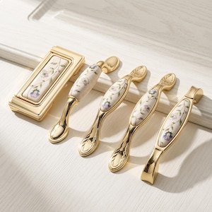 Könnte beinhalten: Ein Set aus goldfarbenen Möbelbeschlägen, bestehend aus einem rechteckigen Knopf und vier geschwungenen Griffen. Jedes Teil ist mit einer weißen Keramikeinlage versehen, die mit einem zarten Blumenmuster in Violett und Grün verziert ist.