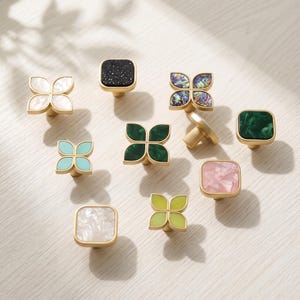 May include: A collection of ten decorative cabinet knobs featuring gold-toned metal bases. The knobs include various shapes such as four-petaled flowers and squares, with inlaid surfaces in white, black glitter, iridescent shell, green, pink, and yellow.