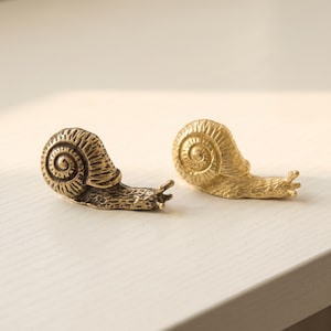 May include: Two small snail figurines crafted from metal. One snail features a dark, antique bronze finish with visible textured details on the shell and body, while the other snail has a bright, polished gold-tone finish with a smooth surface.