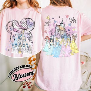 Puede incluir: Camiseta rosa Comfort Colors con un diseño de cuento de hadas. La parte delantera presenta un castillo y un lazo de Minnie Mouse. La parte trasera muestra princesas de Disney frente a un castillo con fuegos artificiales y estrellas. La palabra "Blossom" también es visible.