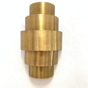Peut inclure: Une applique murale en laiton brossé, présentant un design cylindrique à gradins avec cinq bandes circulaires empilées de largeurs variables, créant un profil symétrique et étagé avec une finition dorée métallique.