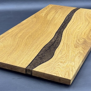 May include: Rectangular wooden serving board with a dark brown epoxy river design. The board is made of light-colored wood with visible grain and a smooth finish. The epoxy river runs down the center, creating a striking contrast.
