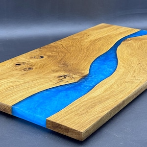 May include: A rectangular wooden serving board with a vibrant blue resin river running through the center. The wood has a natural grain pattern and a warm, golden-brown color. This decorative board is ideal for serving appetizers.