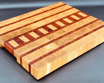 May include: A rectangular wooden cutting board with a light brown and reddish-brown striped pattern. The board is made of end-grain wood, with a series of rectangular blocks and stripes. The board has a smooth surface and is designed for kitchen use.