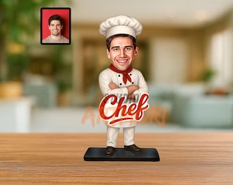 Op de afbeelding: Een beeldje van een chef-kok in een wit uniform, koksmuts en rode halsdoek, met gekruiste armen. Het woord "Chef" is in rode letters op de voorkant geschreven. Een klein ingelijst portret staat op de achtergrond.