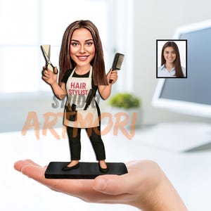 Puede incluir: Una caricatura de una estilista que sostiene tijeras y un peine, con una camiseta negra y un delantal con el texto "HAIR STYLIST". La figura está sobre una base negra. Una foto enmarcada de una mujer está en el fondo.
