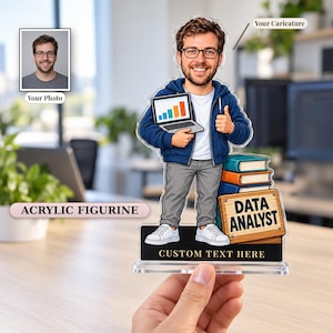 Puede incluir: Una figura de acrílico personalizada con la caricatura de un hombre con gafas, sudadera azul, pantalones grises y zapatillas blancas. La figura sostiene un portátil, está junto a una pila de libros y muestra el texto DATA ANALYST.