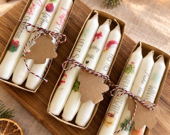 Könnte beinhalten: Drei rechteckige Pappschachteln mit jeweils drei weißen Stabkerzen. Die Kerzen sind mit verschiedenen weihnachtlichen Illustrationen und Texten wie Merry Christmas, Let It Snow und Ho Ho Ho bedruckt. Jede Schachtel ist mit rot-grünem Garn umwickelt.