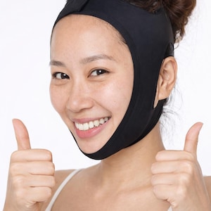 Peut inclure: Une personne porte une sangle noire amincissante pour le visage, souriante et faisant un signe de pouce levé. La sangle couvre le menton et la mâchoire, avec une bande sur le front. La personne porte un débardeur blanc.