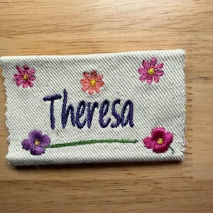 May include: Embroidered name tag with the name "Theresa" in purple script. The tag is decorated with embroidered pink and purple flowers and a green stem. The fabric is a light beige color.