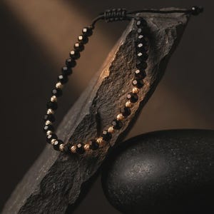 May include: A bracelet featuring alternating black faceted beads and small metallic gold textured spheres. The bracelet is constructed with a black cord and includes an adjustable sliding knot closure mechanism for a secure fit.