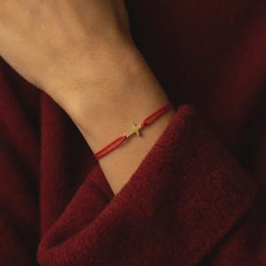 May include: A delicate bracelet featuring a thin, vibrant red cord and a small, polished gold-toned cross charm centered on the wrist. The minimalist design consists of two strands of red cord that hold the metallic cross element securely in place.
