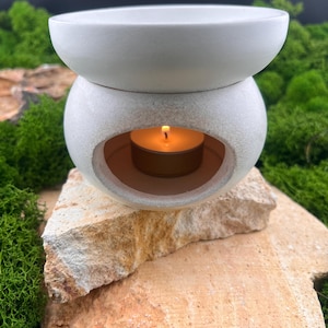Könnte beinhalten: Ein mattweißer Keramik-Ölbrenner mit abgerundeter Basis und einer breiten Schale oben. Ein kleines, brennendes Teelicht mit warmer orangefarbener Flamme ist in der zentralen Öffnung sichtbar, platziert auf einem strukturierten, hellen Stein.
