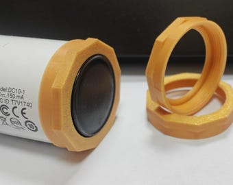 May include: A white cylindrical device with a black speaker and orange hexagonal trim. Two additional orange hexagonal rings are next to the device. The device has the text "Model: DC10-1 5 V..." and "FCC ID: T7V1740".