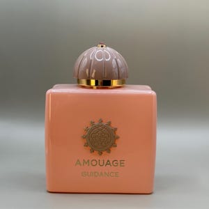 May include: A peach-colored perfume bottle with a rounded top and gold accents. The bottle has the words "AMOUAGE GUIDANCE" printed on the front. The top is a light brown color with a small pink gem on top.