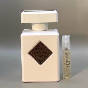Könnte beinhalten: Eine weiße Parfümflasche mit rechteckiger Form und quadratischem Deckel. Ein braunes rautenförmiges Emblem befindet sich auf der Flasche. Eine kleine Flasche mit der Aufschrift "Initio Paragon 3ml" daneben.