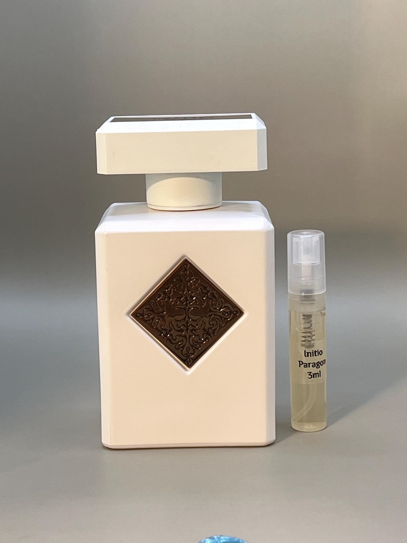 Peut inclure: Un flacon de parfum blanc de forme rectangulaire avec un bouchon carr&eacute;. Un embl&egrave;me en forme de losange marron est centr&eacute; sur le flacon. Un petit flacon avec l'inscription "Initio Paragon 3ml" est &agrave; c&ocirc;t&eacute;.