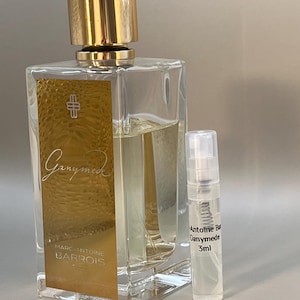 Puede incluir: Un frasco de perfume de vidrio transparente con una tapa dorada y un frasco de muestra más pequeño. El frasco más grande tiene una etiqueta dorada con la palabra "Ganymede" y la marca "Marc-Antoine Barrois". El frasco de muestra dice "Antoine Barrois Ganymede 3ml".