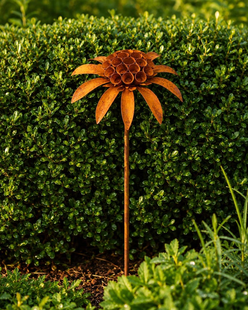 K&ouml;nnte beinhalten: Ein rostfarbener Gartenstecker aus Metall in Form einer Blume. Der Bl&uuml;tenkopf besteht aus einer zentralen Ansammlung kleiner, kreisf&ouml;rmiger, hohler R&ouml;hren, die von einem Kranz aus langen, spitzen Bl&uuml;tenbl&auml;ttern umgeben sind. Ein d&uuml;nner, gerader Stab.