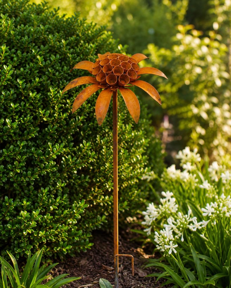 K&ouml;nnte beinhalten: Ein rostfarbener Gartenstecker aus Metall in Blumenform. Die Bl&uuml;te besteht aus einem zentralen B&uuml;ndel kleiner, hohler zylindrischer R&ouml;hren, umgeben von einer Reihe langer, spitzer Bl&uuml;tenbl&auml;tter. Das Ganze wird von einem d&uuml;nnen Metallstab getragen.