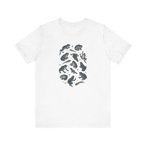 May include: A white short-sleeved t-shirt featuring a central graphic print of various dark gray frogs and tadpoles arranged in an oval cluster, interspersed with small four-pointed star shapes across the front of the garment.