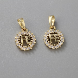 May include: Two gold-colored pendants, each featuring the letters "FE" in the center, surrounded by a halo of small, clear crystals. The pendants have a loop at the top for a chain. The background is a solid gray.