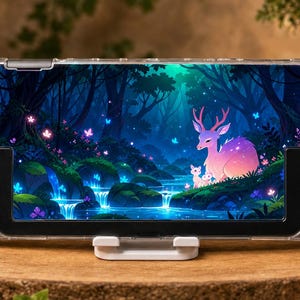 May include: A handheld gaming console featuring a clear protective case and a vibrant skin depicting a glowing forest scene with a deer, a fawn, and a waterfall. The device includes black joysticks, directional buttons, and action buttons.
