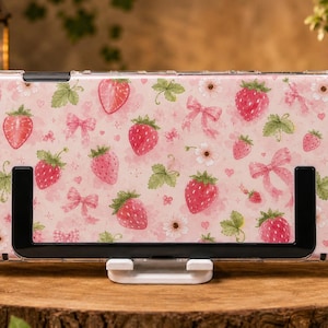 Könnte beinhalten: Eine tragbare Spielkonsole mit einer hellrosa Schutzhülle, die mit einem wiederkehrenden Muster aus roten Erdbeeren, grünen Blättern, kleinen weißen Blüten und hellrosa Schleifen verziert ist. Das Gerät hat schwarze Tasten und Joysticks.