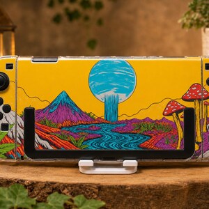 May include: A handheld gaming console featuring a vibrant yellow skin with a colorful landscape illustration. The design includes a blue moon, a flowing river, purple mountains, and red-capped mushrooms. The console has black buttons and joysticks.