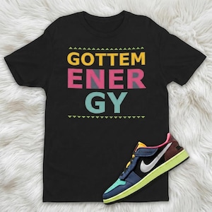 May include: Black t-shirt with the words "GOTTEM ENERGY" in yellow, pink, gray, and teal. A colorful sneaker with blue, green, yellow, and white accents is next to the shirt.