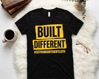 May include: Black t-shirt with the words "BUILT DIFFERENT" in yellow block letters. Below the text is the hashtag "#CUTFROMADIFFERENTCLOTH". The shirt is on a white fluffy surface, with a cup of coffee, glasses, and books in the background.